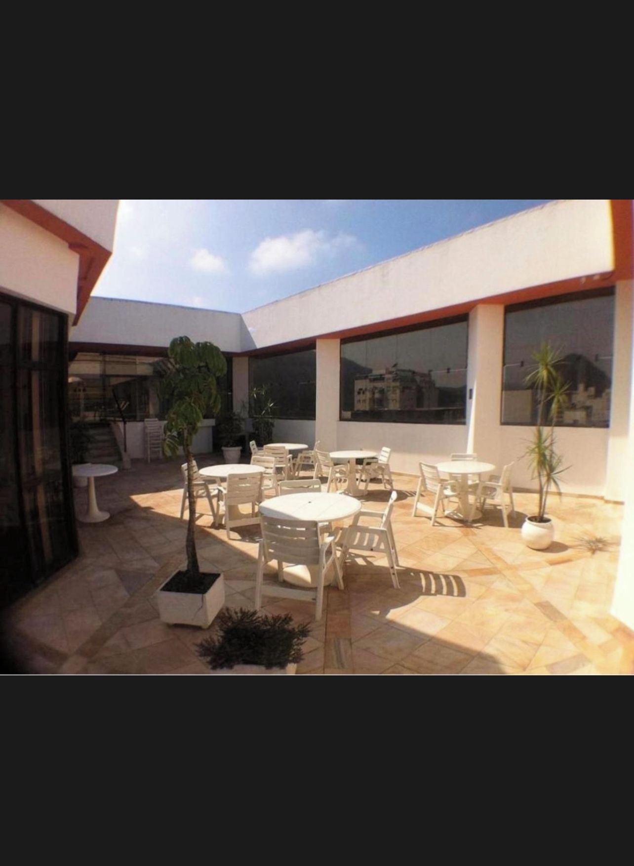 Un espace extérieur aménagé en terrasse, avec plusieurs tables et chaises rondes, des plantes en pot et de grandes fenêtres offrant une vue sur les bâtiments environnants.