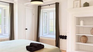 A bedroom with a bed, a hanging light fixture, a radiator, a window with curtains, and a white shelving unit with decorative items.