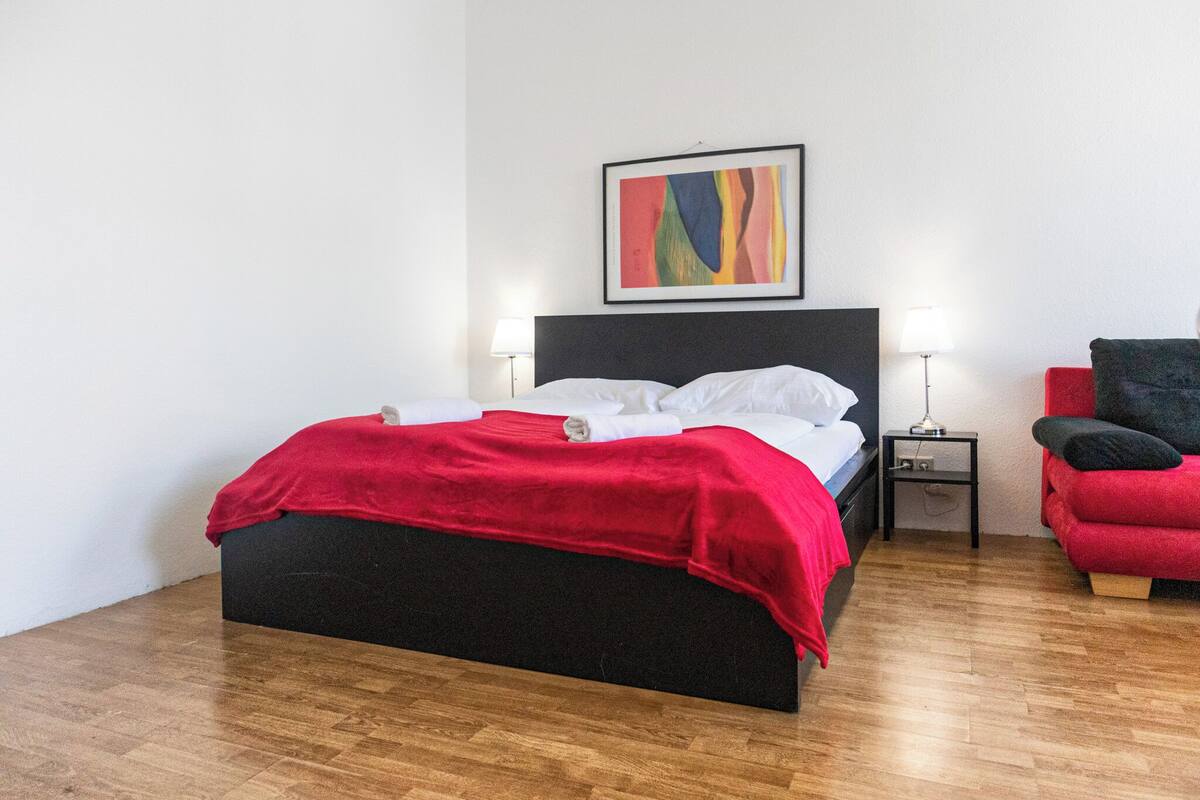 A modern hotel room with a black bed, red accents, a colorful abstract painting, and a red armchair.
