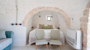 Quarto com arco de pedra, cama com duas almofadas, mesa de cabeceira, uma cadeira e radiador.