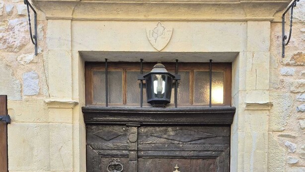 Uma porta dupla de madeira com um bate-metal e uma pequena placa, em um edifício de pedra com fachada simples e sem adornos.