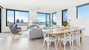 Un salon moderne avec un canapé gris, deux fauteuils, une table à manger en bois avec des chaises blanches et de grandes fenêtres offrant une vue sur le ciel et les arbres.