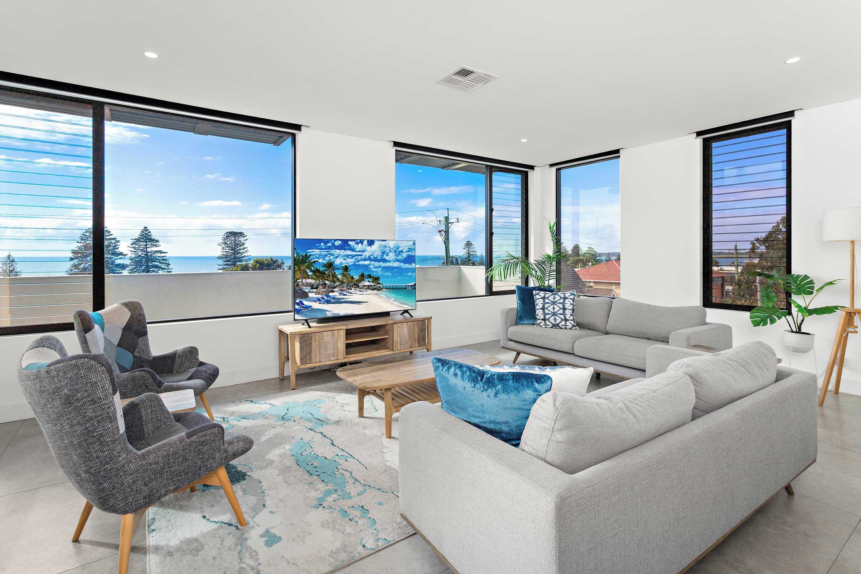 A modern living room with large windows, a grey sofa, a grey armchair, a coffee table, and a TV on a wooden stand.