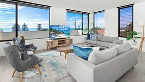 Un salon moderne avec de grandes fenêtres, un canapé gris, un fauteuil gris, une table basse et un téléviseur posé sur un meuble en bois.