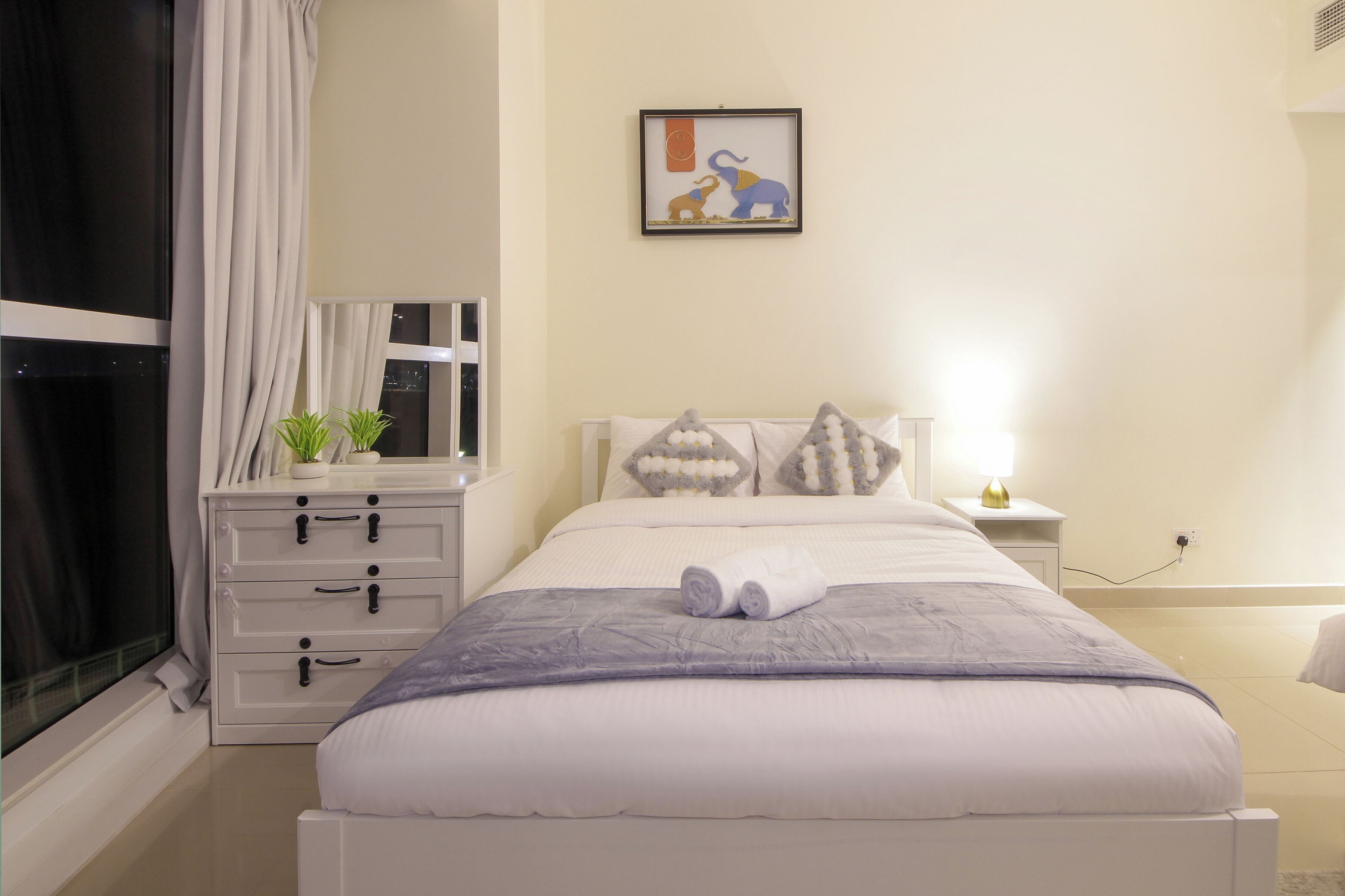 A neatly made bed with a white comforter and pillows, a bedside table with a lamp, and a window with curtains.