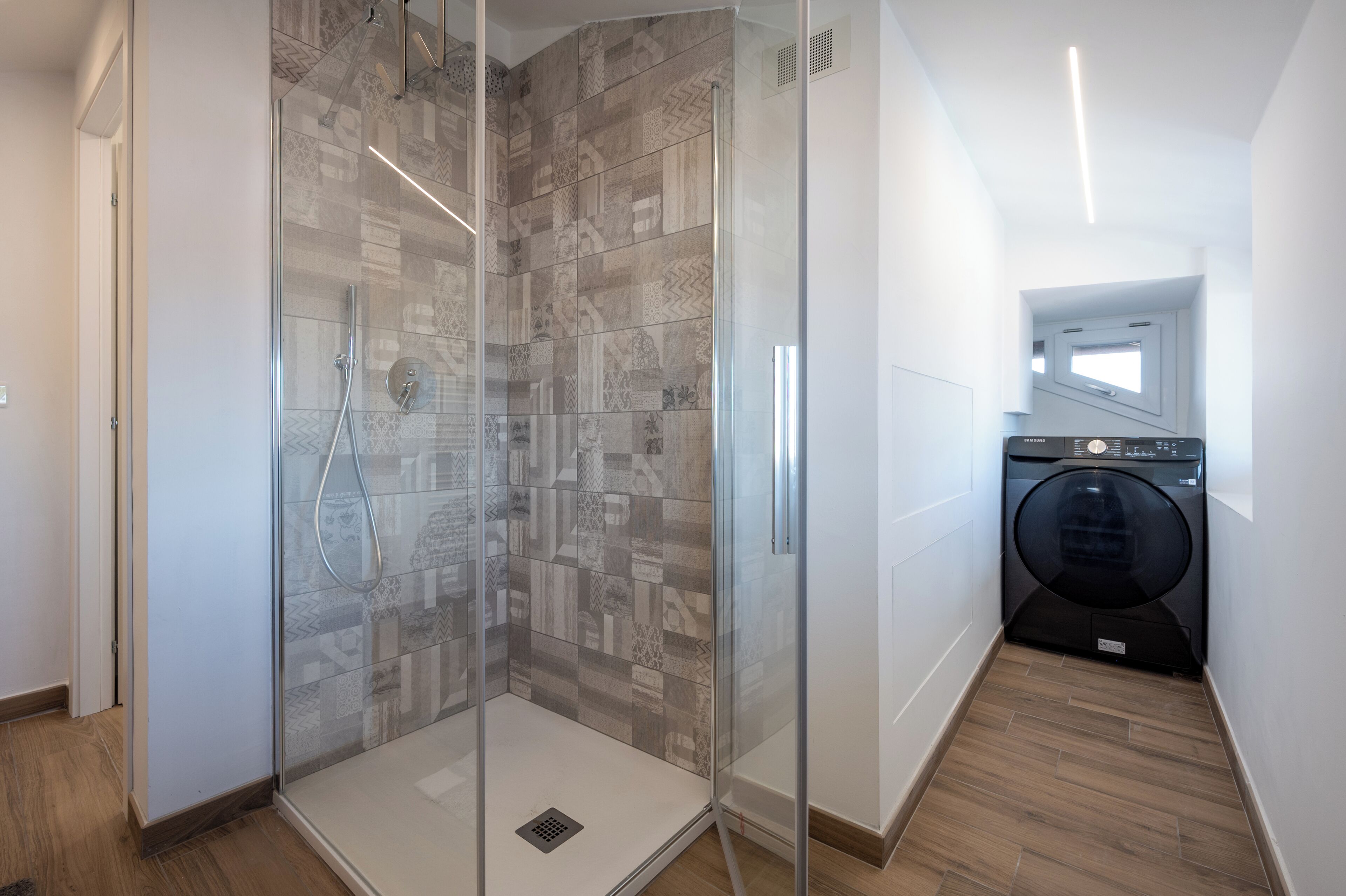 Un baño moderno con cabina de ducha de vidrio, lavadora integrada y una ventana pequeña.