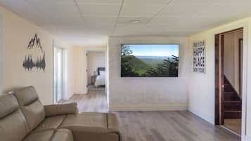 A modern living room with a large flat-screen TV mounted on a white wall, a beige sofa, and a staircase leading to another room.