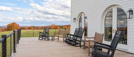 A wooden deck with outdoor furniture, a white building with arched windows, and a view of autumn foliage.