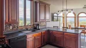 Una cocina con armarios de madera oscura, una encimera negra y un lavavajillas. Amplias ventanas ofrecen vistas al exterior.