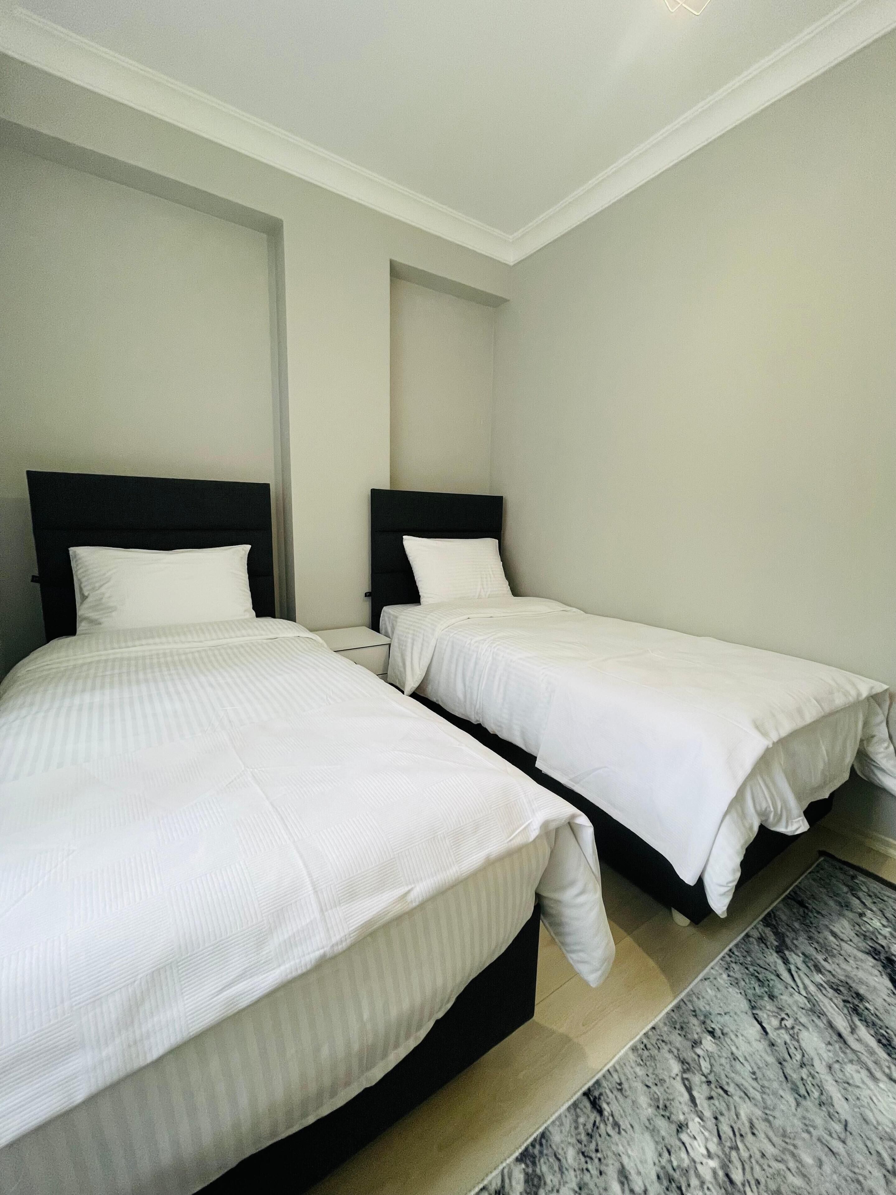 Ein Hotelzimmer mit zwei Einzelbetten, jeweils mit schwarzem Kopfteil und weißer Bettwäsche. Der Raum hat einen Marmorboden und eine schlichte weiße Wand.