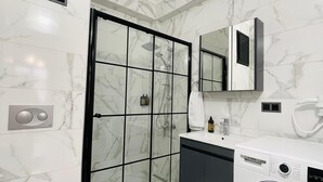 A modern bathroom with a marble floor, a glass shower enclosure, a built-in washing machine, and a large mirror above the sink.