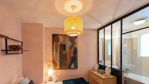 A modern bedroom with a bed, bedside table, lamp, abstract artwork, and a large window with a view of the bathroom.