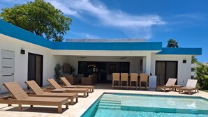 Un espace moderne aménagé au bord de la piscine, avec des chaises longues, un coin repas et une cuisine visible à travers une porte ouverte.
