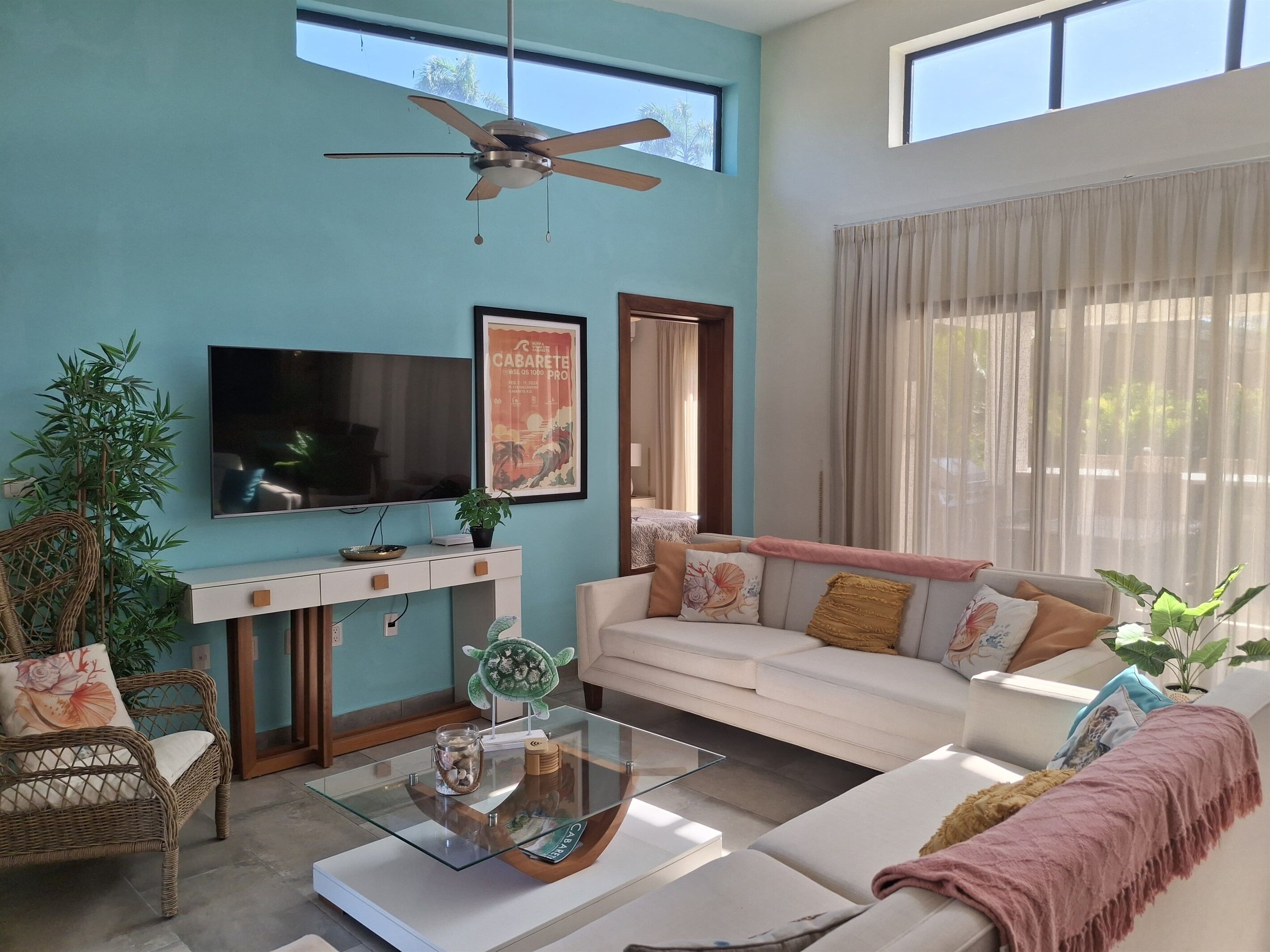 Ein modernes Wohnzimmer mit Flachbildfernseher, Deckenventilator, Sofa, Couchtisch und einem großen Fenster mit Vorhängen.