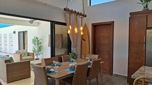 Un coin repas avec une table en bois dressée pour six personnes, entourée de chaises en osier. Une planche de surf est appuyée contre le mur, ainsi qu’un meuble en bois et un ventilateur de plafond.