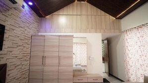 A modern bedroom with a stone-textured wall, a wooden wardrobe, a bed with floral curtains, and a bathroom visible through a glass door. - Kisan Farm House | Serene 2BR Villa with Private Pool & Garden, Hillside Stay (Rajsamand)