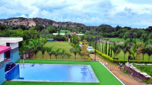 A residential area with a swimming pool, green lawns, and a hilly landscape in the background. - Kisan Farm House | Serene 2BR Villa with Private Pool & Garden, Hillside Stay (Rajsamand)