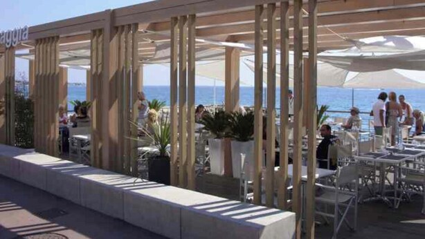 An outdoor seating area with tables and chairs, a pergola structure, and a view of the sea.