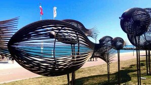 A series of abstract metal sculptures resembling fish on a grassy area with a clear blue sky and a beach in the background.