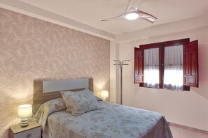 Une chambre à coucher comprenant un lit, des tables de chevet, une lampe, un ventilateur de plafond et une fenêtre avec des rideaux.