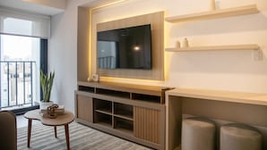 A modern living room with a flat-screen TV mounted on a wooden wall, a small table with books, a potted plant, and a view of the city through a large window. - w Sunlit 1BR w Private Balcony in Miraflores (Lima)