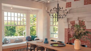 A dining area with a wooden table, chairs, and a bench with cushions. There are large windows with a view of greenery outside. - Villa des Rêves: Luxury in Villers, Pool, Jacuzzi & Padel court, A/C, close to Deauville (Villers-sur-Mer)