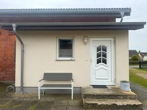 Un edifici petit amb una porta blanca, una finestra i una banca a l'exterior.