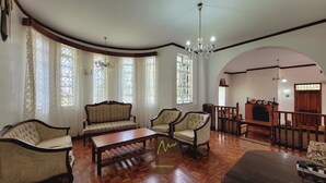 A spacious living room with a wooden floor, a sofa set, armchairs, and a coffee table. There are large windows with curtains, a chandelier, and a fireplace. - Tina's ngong hills cottage - country living (Kajiado County)