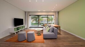 Ein modernes Wohnzimmer mit Flachbildfernseher, grauem Sofa, Poufs, einem kleinen runden Tisch und einem Esstisch mit Stühlen.