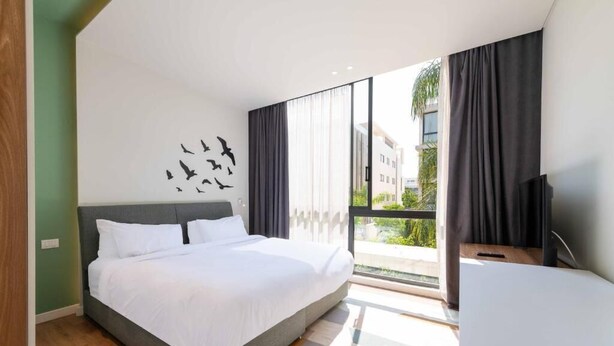 Ein modernes Hotelzimmer mit einem großen Bett, einem Flachbildfernseher und einem Balkon mit Blick auf Gebäude und Grünflächen.