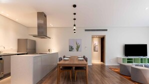 Eine moderne Küche mit Kochinsel, ein Essbereich mit Holztisch und -stühlen sowie ein Wohnbereich mit Flachbildfernseher und grünen Regalen.