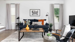 A modern living room with a dark blue sofa, a wooden coffee table, and a black armchair. There is a framed picture on the wall, a plant on the sofa, and a TV on the right. - Modern 4BR Retreat for 12, Near Downtown Nash! (Nashville)