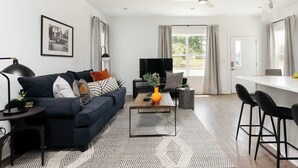 A modern living room with a dark blue sofa, a coffee table, and a TV. There is a dining area with a white table and black chairs. The room has a large window with curtains and a ceiling fan. - Modern 4BR Retreat for 12, Near Downtown Nash! (Nashville)