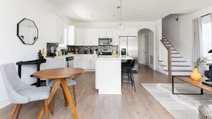 A modern kitchen with white cabinetry, a central island, and a dining area with a round wooden table and grey chairs. - Modern 4BR Retreat for 12, Near Downtown Nash! (Nashville)
