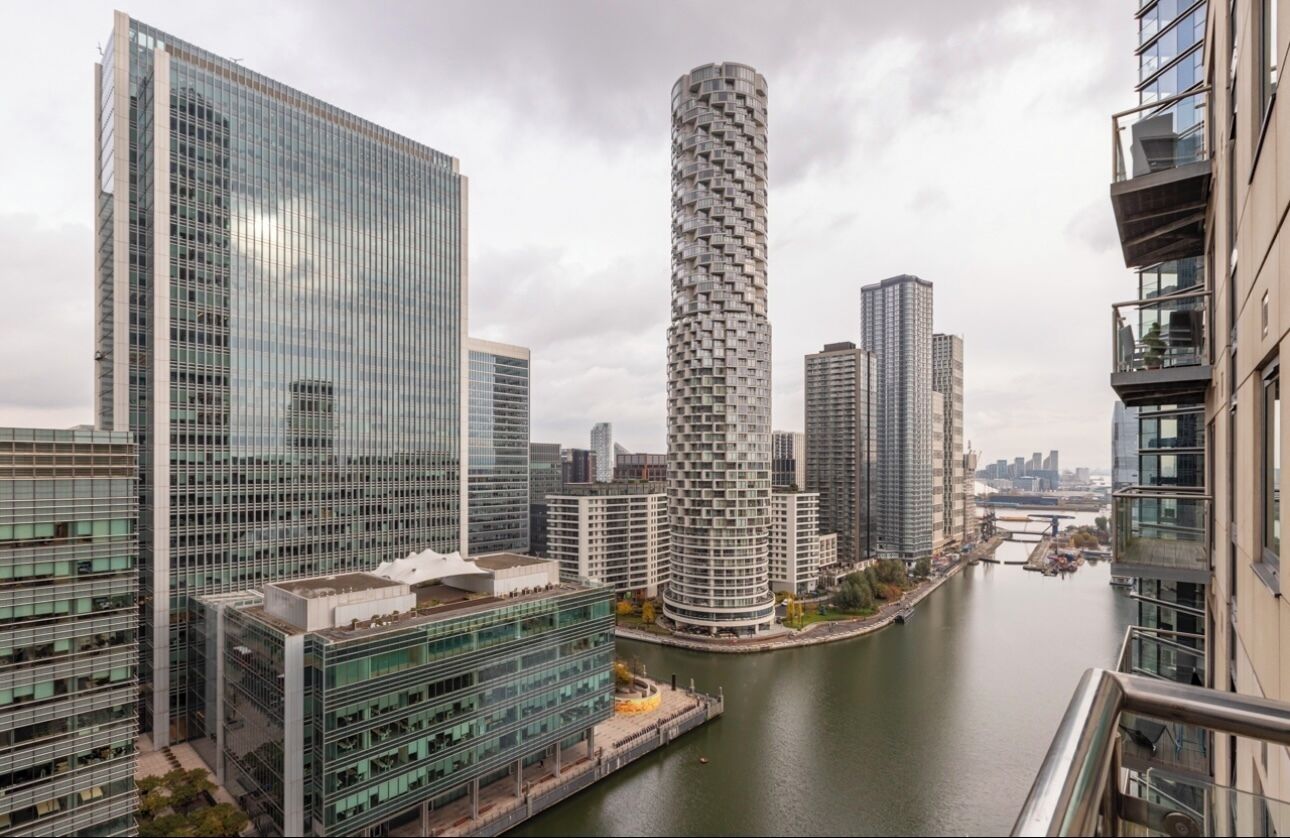 A modern high-rise building with a unique cylindrical design, surrounded by other contemporary skyscrapers and a river.