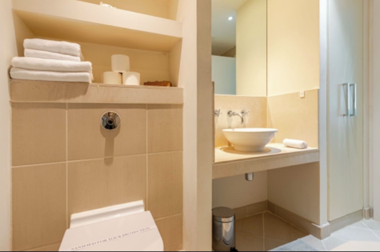 Casa de banho moderna com lavatório branco, sanita e espelho. Toalhas empilhadas numa estante e um cesto de lixo sob o lavatório.