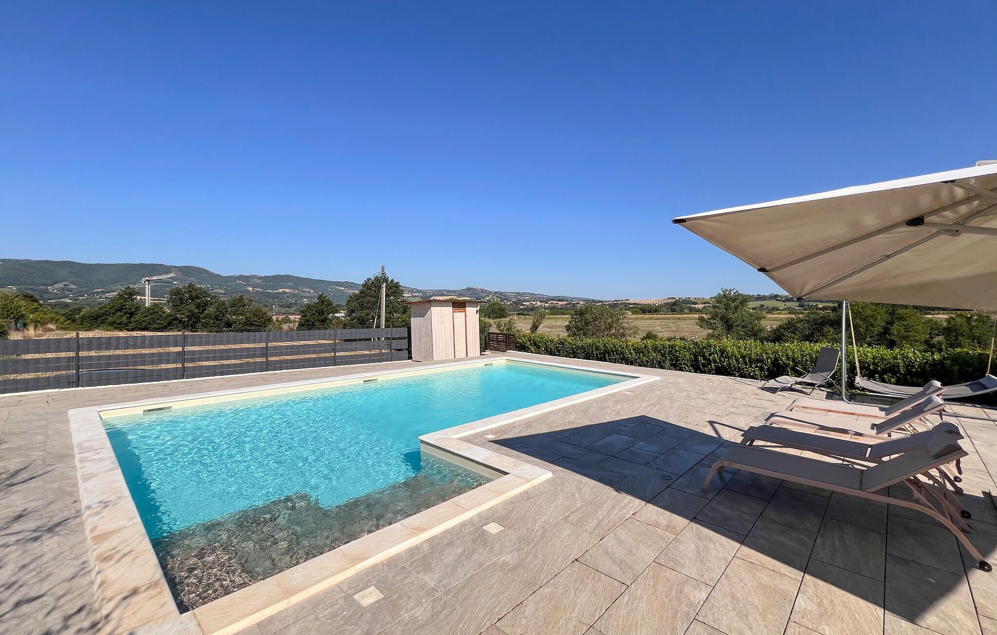 A rectangular swimming pool with a clear blue water, surrounded by a tiled patio area. There are lounge chairs and a large umbrella providing shade.
