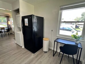 A compact kitchen with a black refrigerator, a small trash bin, a window with a view of the street, and a dining area with chairs and a table.