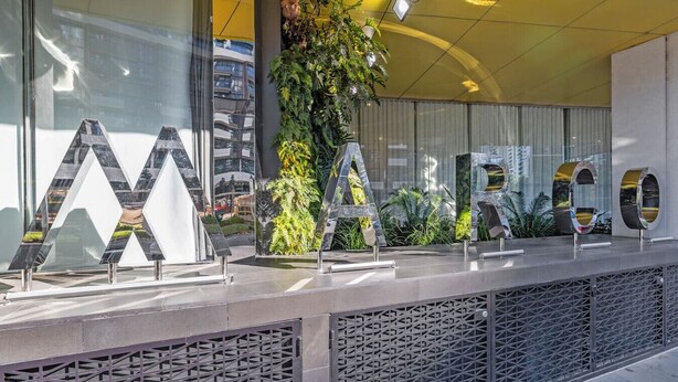 Fachada moderna de un edificio con letras metálicas grandes que deletrean "MARCO" sobre una superficie reflectante, rodeada de follaje y una valla de metal geométrica.