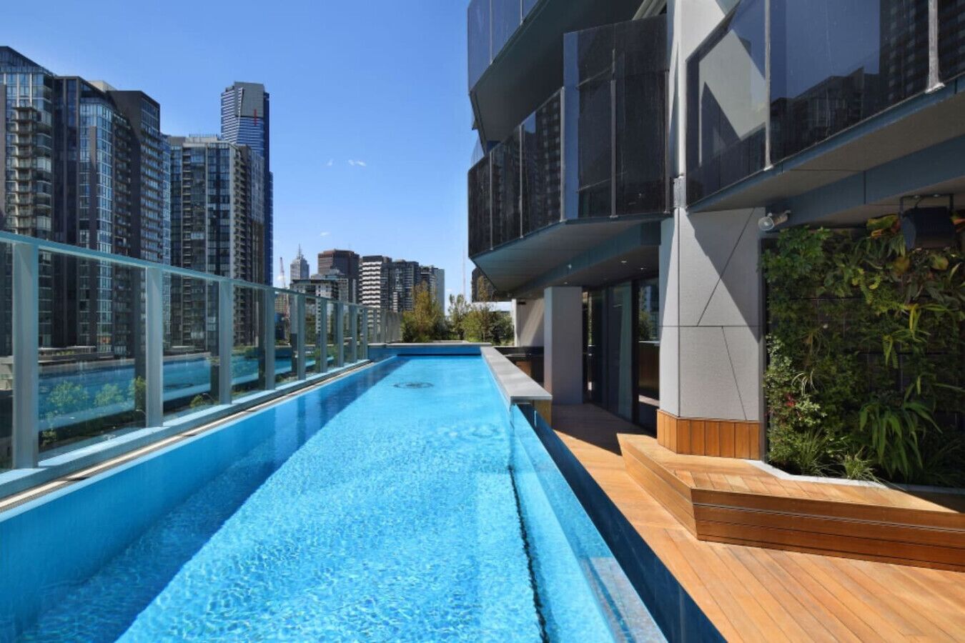 A rooftop pool with a glass barrier, surrounded by high-rise buildings and a wooden deck.