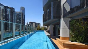 Una piscina en la azotea con barandilla de vidrio, rodeada de rascacielos y una terraza de madera.
