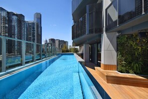 Una piscina en la azotea con barandilla de vidrio, rodeada de rascacielos y una terraza de madera.