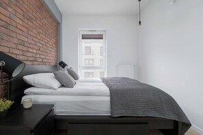 Quarto com parede de tijolos, cama com roupa de cama branca, luminária de cabeceira e vista para edifícios pela janela.