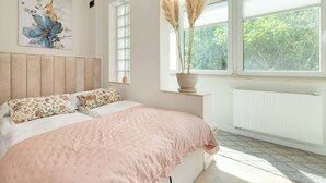 Un dormitorio con cama, ventana con vistas a la vegetación, radiador y un cuadro floral en la pared.