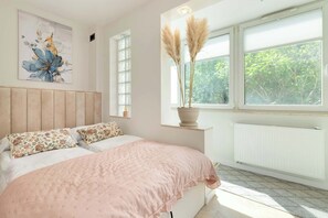 A bedroom with a bed, a window with a view of greenery, a radiator, and a wall with a floral painting.