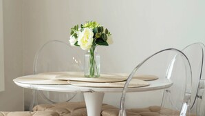 Una mesa redonda de comedor con un florero de vidrio transparente, sillas traslúcidas con asientos acolchados y manteles individuales tejidos.