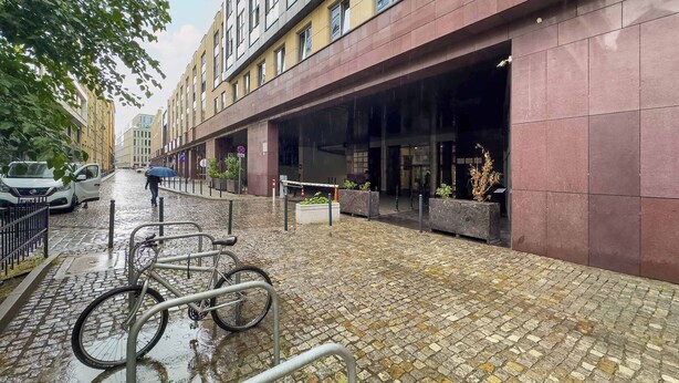 Une rue pavée et mouillée, avec un support à vélos, un piéton et un bâtiment doté d’une grande entrée.