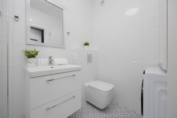 Un baño moderno con azulejos blancos, inodoro, lavabo con espejo y una planta pequeña en la encimera.