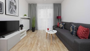 Una sala de estar moderna con un televisor de pantalla plana, una mesita blanca, un sofá gris con almohadones decorativos, una planta en maceta y un ventanal con cortinas de gasa.
