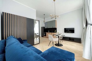 Una sala moderna con un sofá azul, un comedor con una mesa pequeña y sillas, una cocina al fondo y un televisor montado en la pared.